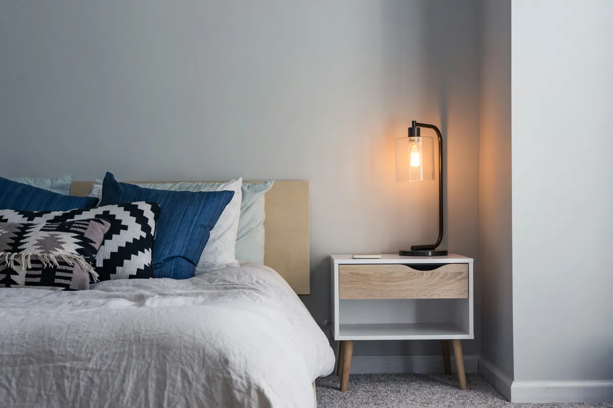 A furnished private bedroom with a made bed, pillows and a bedside lamp
